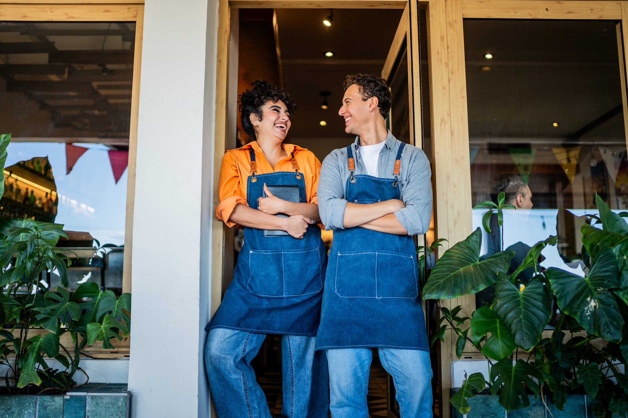 Two restaurant owners in denim aprons laughing in the doorway of their café